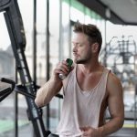 Muscular young sportsman in casual sportswear drinking fresh energy beverage near window while resting after intense workout in modern fitness club
